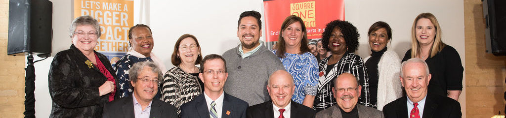 Thirteen adults, diverse in age and background, smiling together in a group photo indoors.