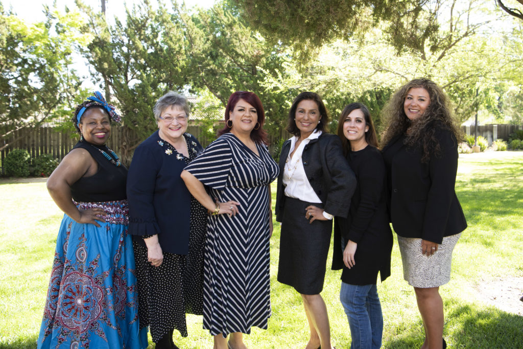 Six women pose and smile together outdoors on a sunny day, with trees in the background.