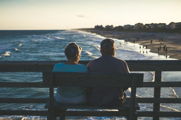 Two people sit on a bench on a pier overlooking the ocean.