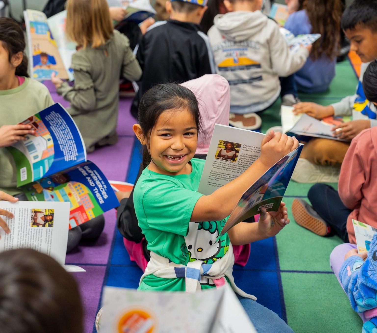 Smiling girl holding a book, sitting with other children reading on a colorful classroom rug.