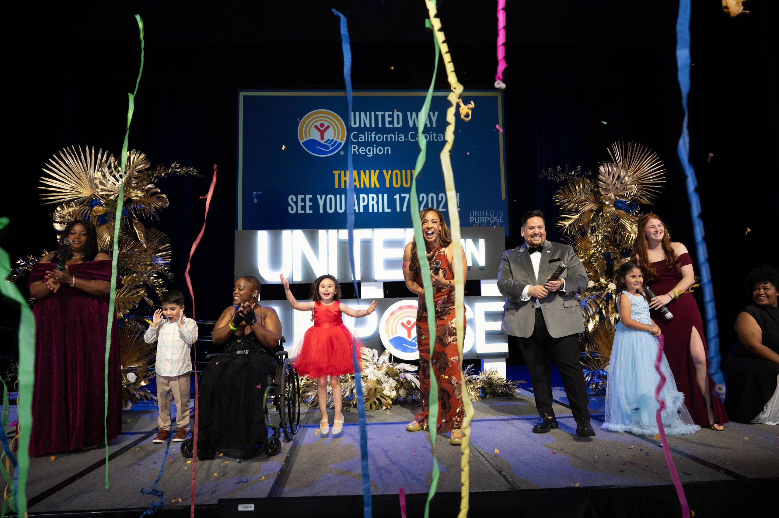 A diverse group of adults and children, some in formal attire, celebrate on stage with confetti streamers. The backdrop reads UNITED WAY CALIFORNIA CAPITAL REGION and SEE YOU APRIL 17, 2024.