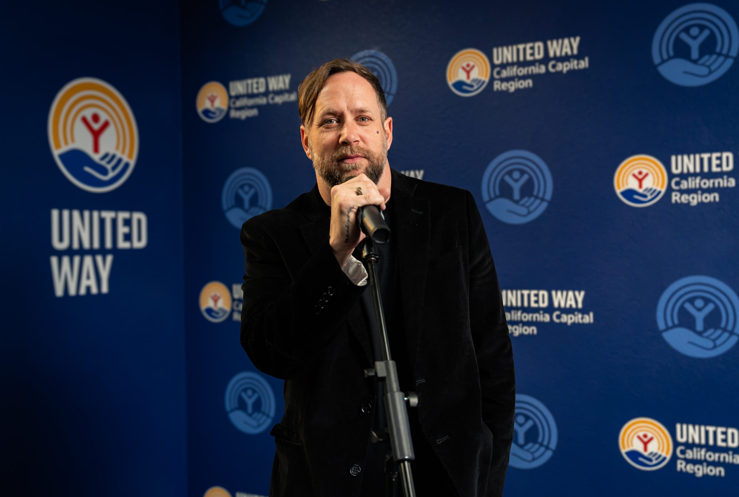 Man in a black jacket speaks at a microphone at a United Way event backdrop.